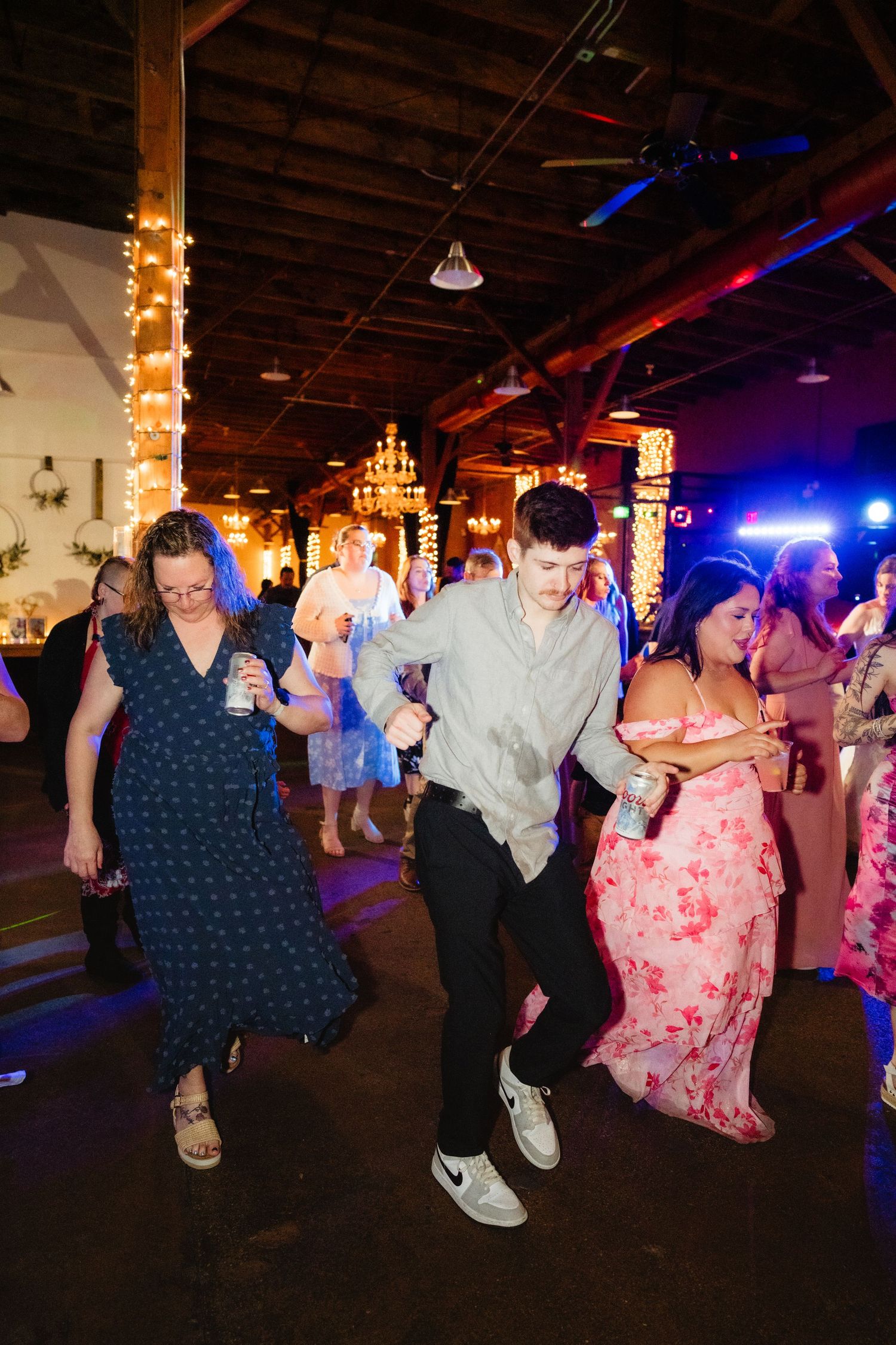 Guests dancing energetically at a wedding reception in a warmly lit rustic venue with string lights.
