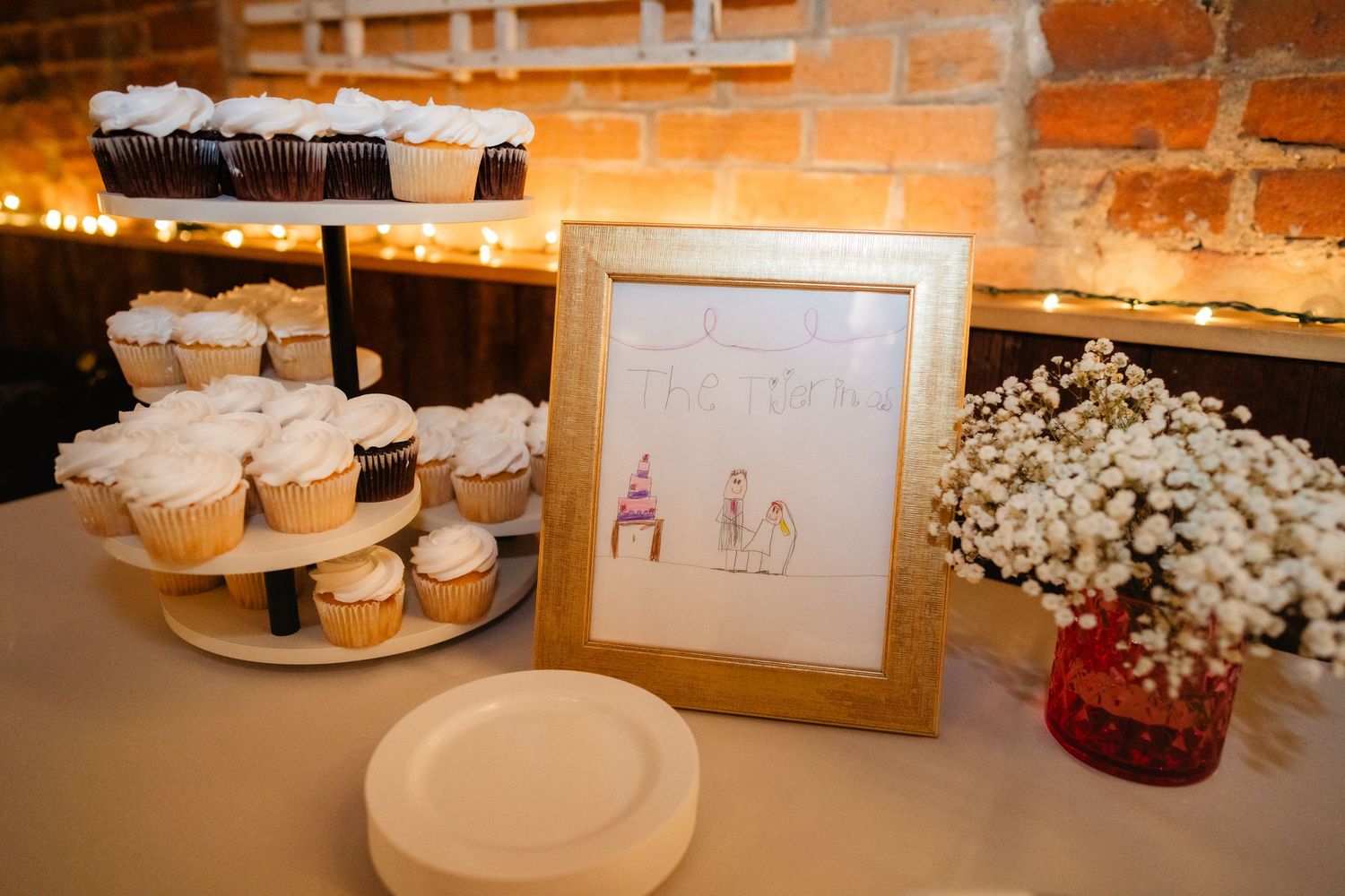 Wedding dessert table with white frosted cupcakes on tiered stand, framed couple illustration, and baby's breath floral arrangement.
