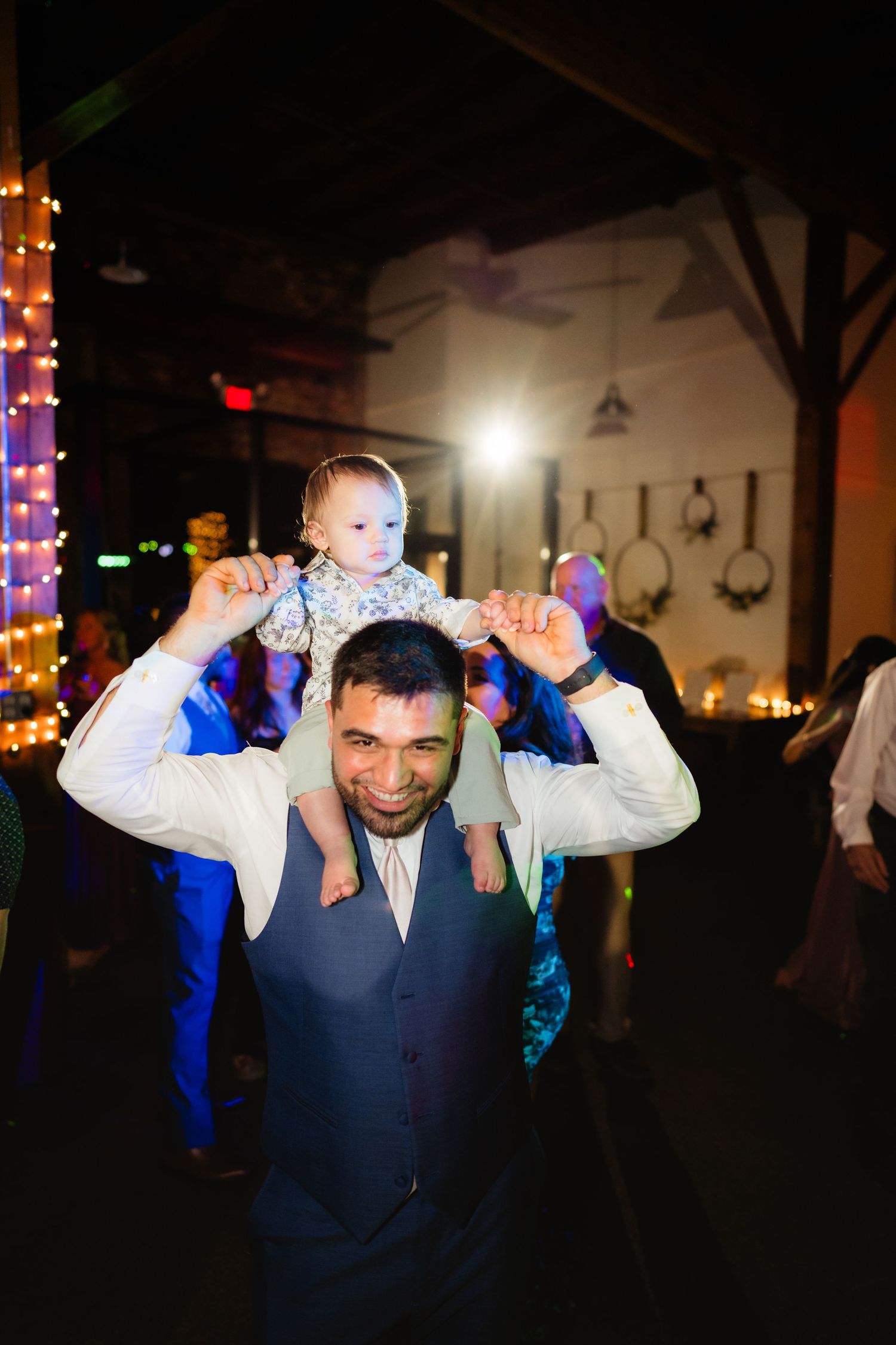 A smiling man in a vest carries a baby on his shoulders while dancing at a wedding reception.