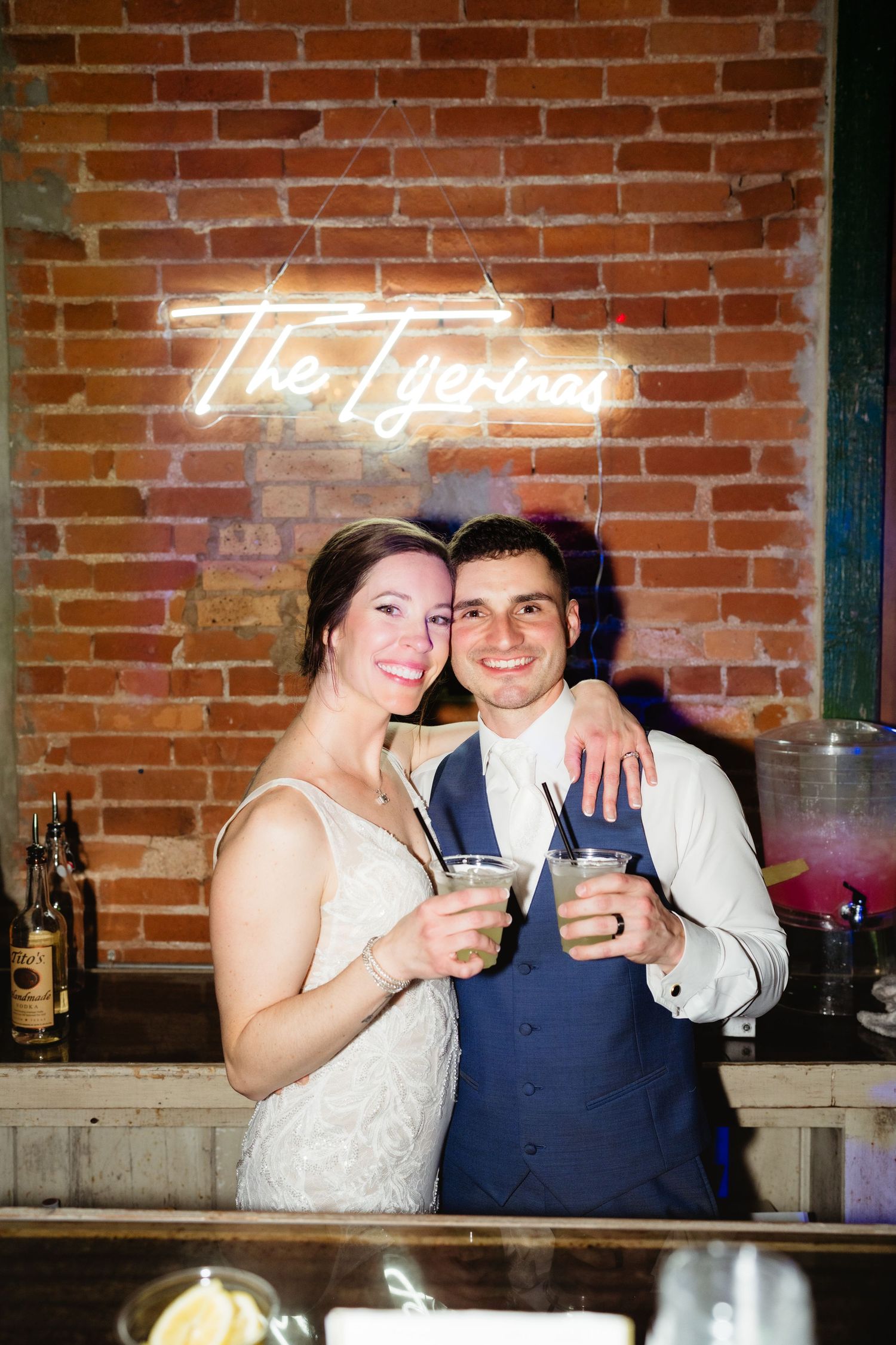 Newlywed couple smiling and toasting drinks at a bar beneath a glowing neon sign at their wedding reception.