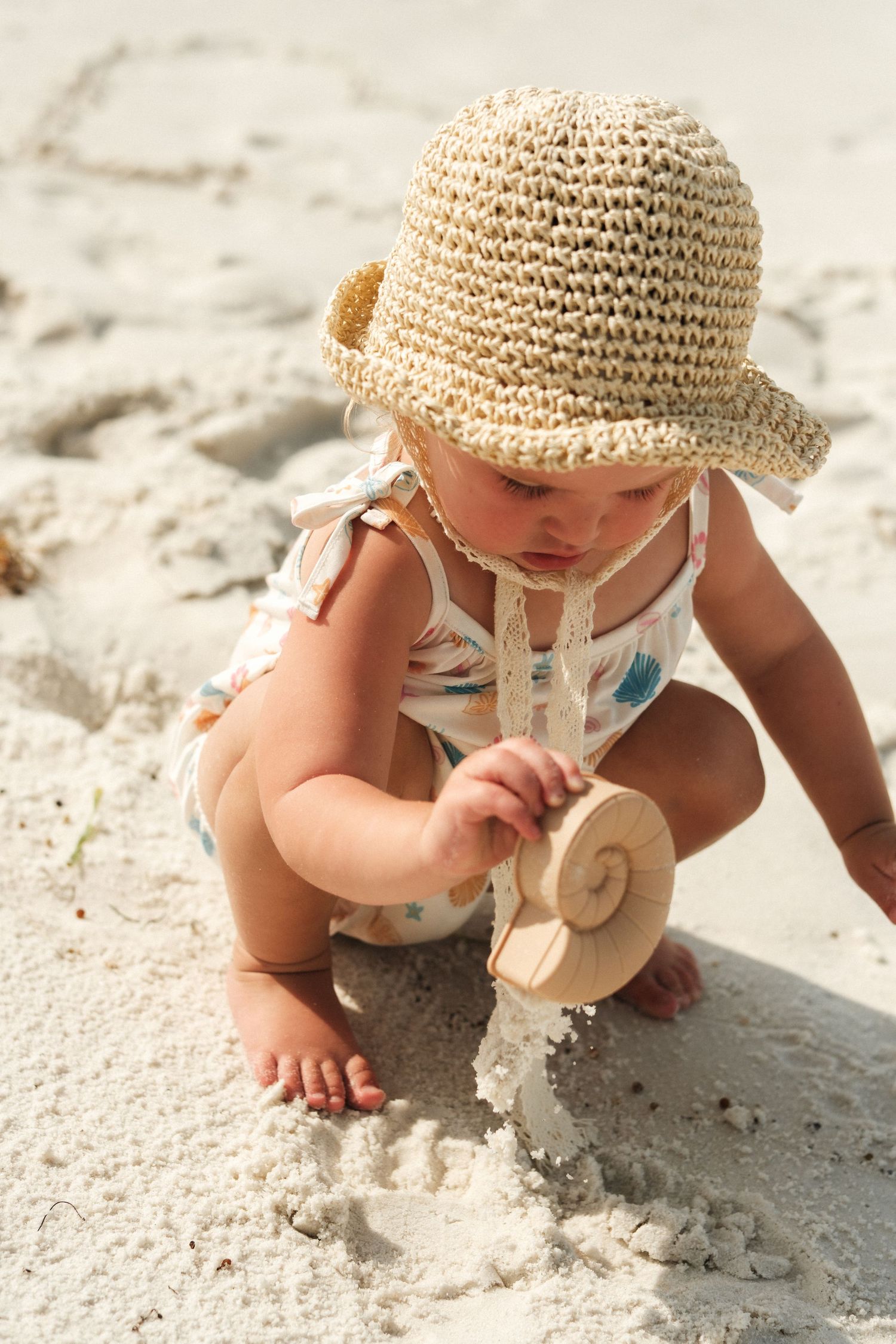 Baby in sunhat playing with seashell in sand on beach.