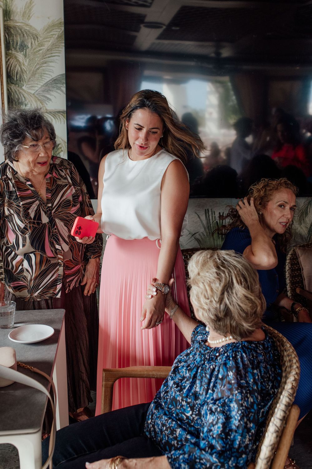 Woman in white top and pink skirt greets elderly guests at an indoor celebration.