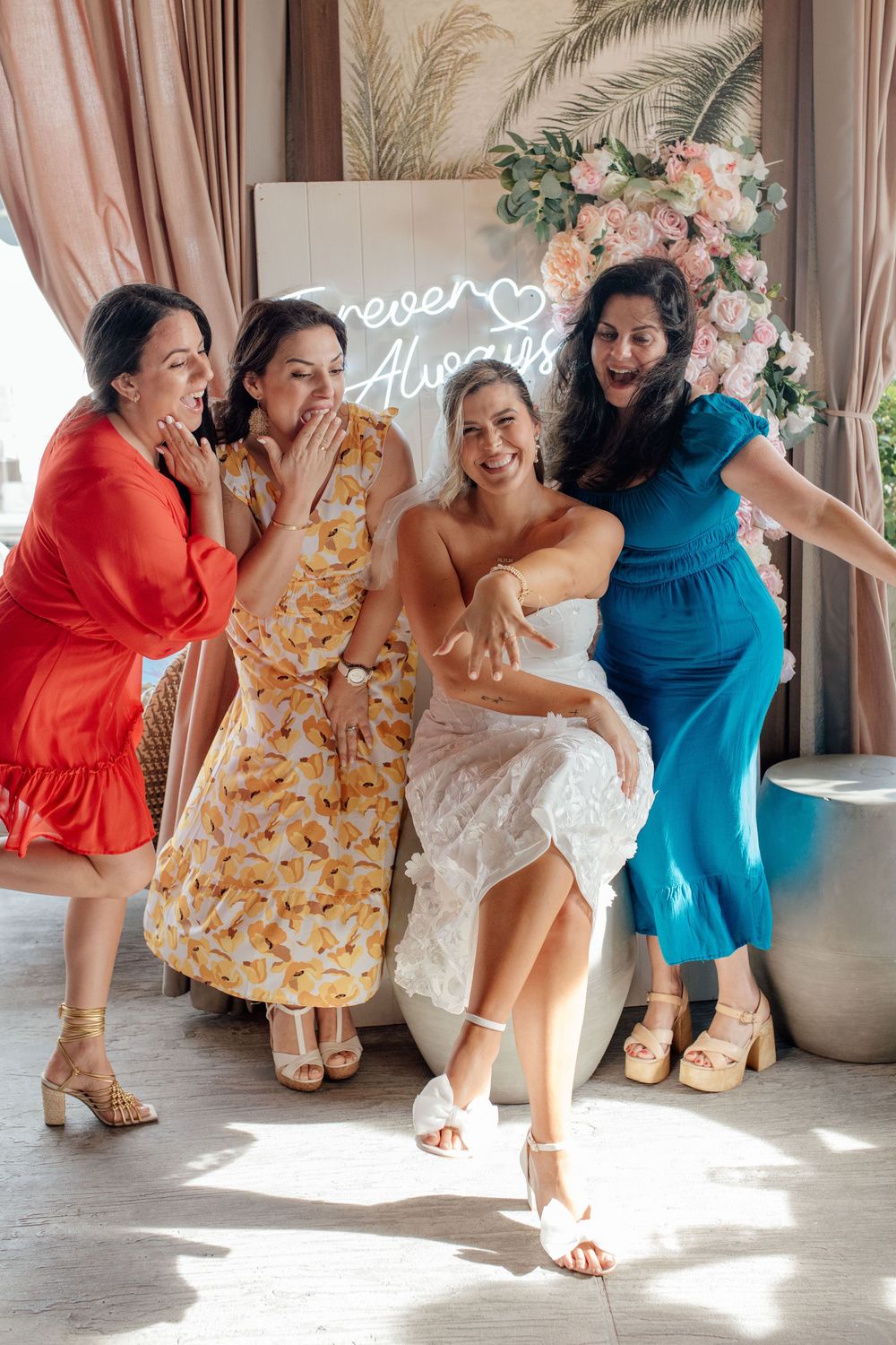 Four joyful women celebrate with a bride in a white dress in front of a Forever Always neon sign.