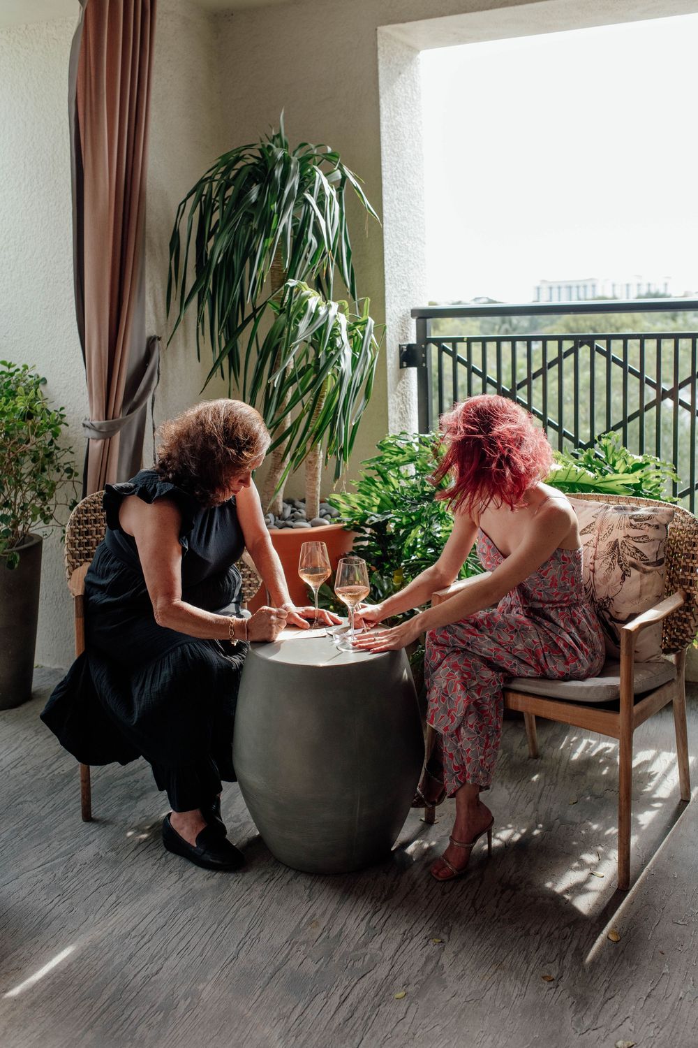 Two women sharing wine on sunny balcony surrounded by lush tropical plants.