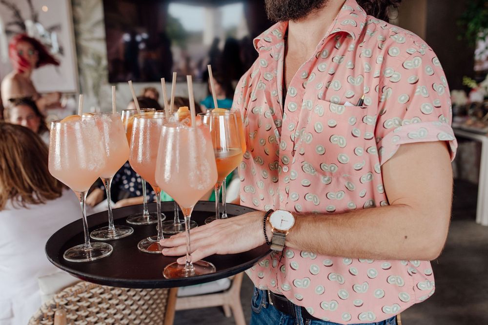 Server in pink watermelon shirt carrying tray of champagne cocktails at wedding reception.