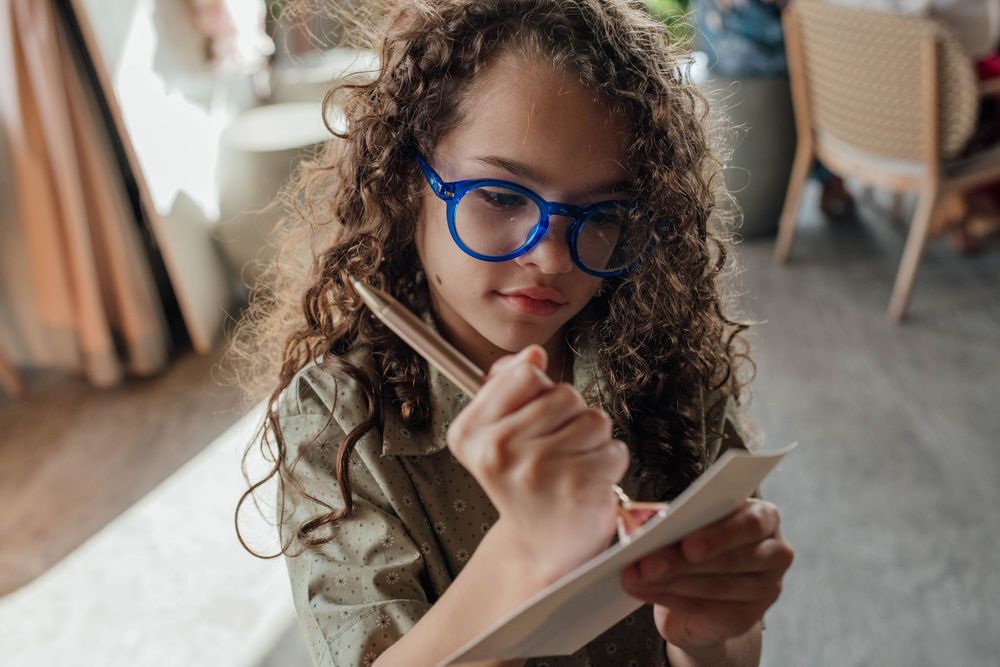 Young girl with curly hair and blue glasses writing in a small notebook with a pencil.