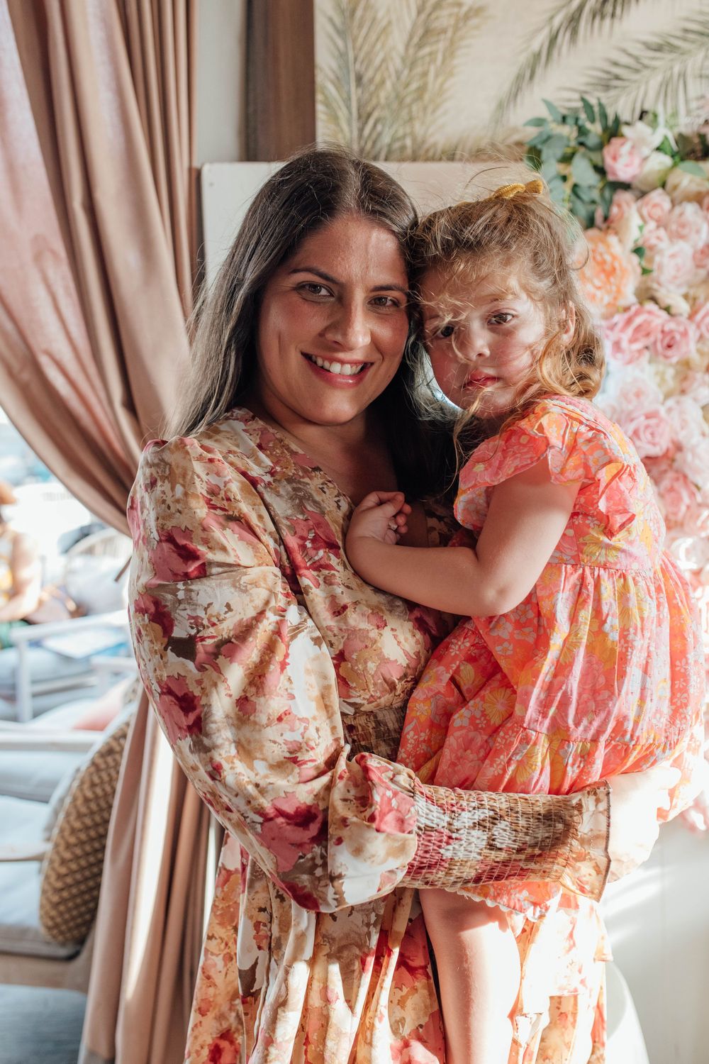 Smiling woman in floral dress holding a young girl in orange dress at a wedding event.