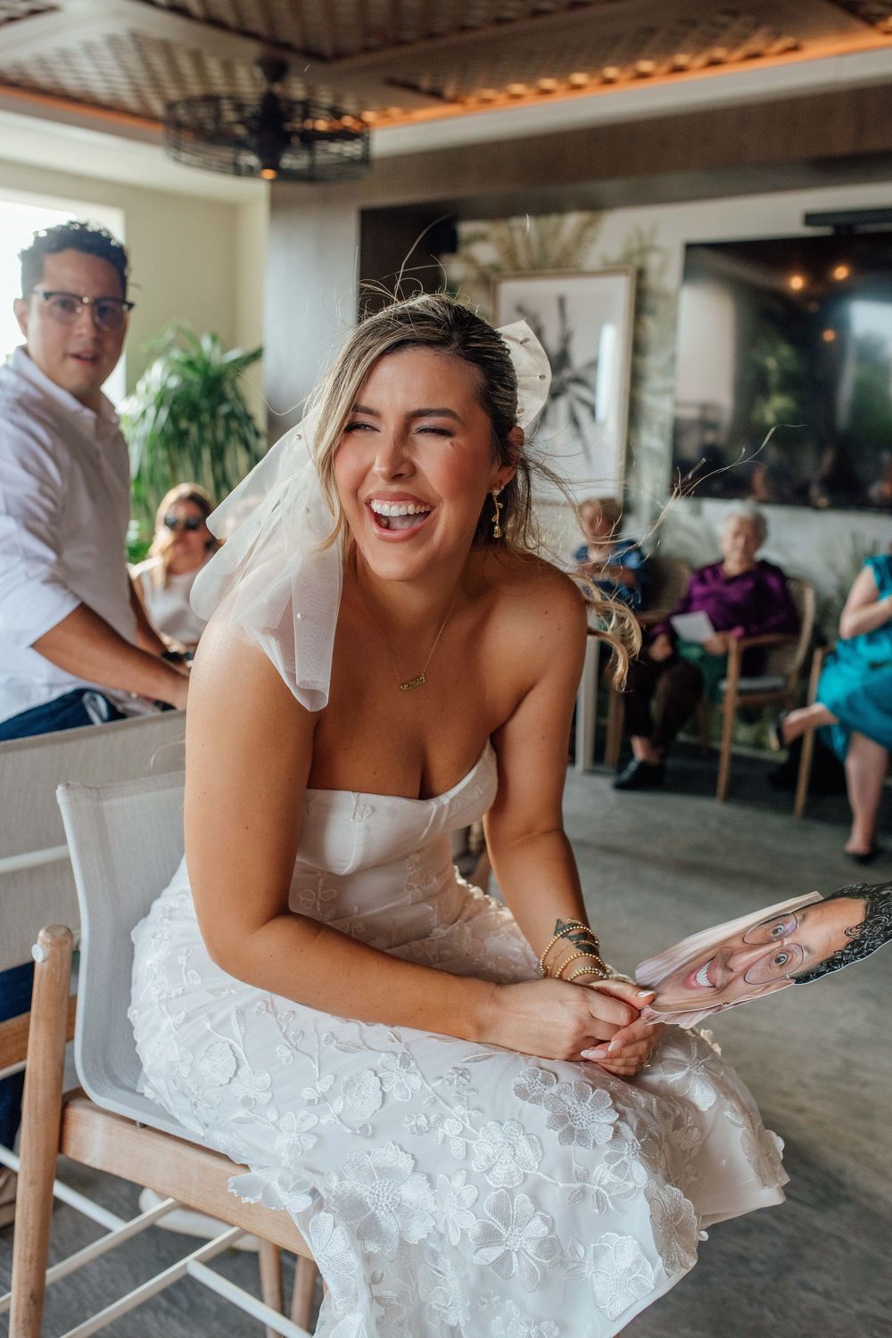 Joyful bride in white strapless dress laughing while holding fan at wedding reception.