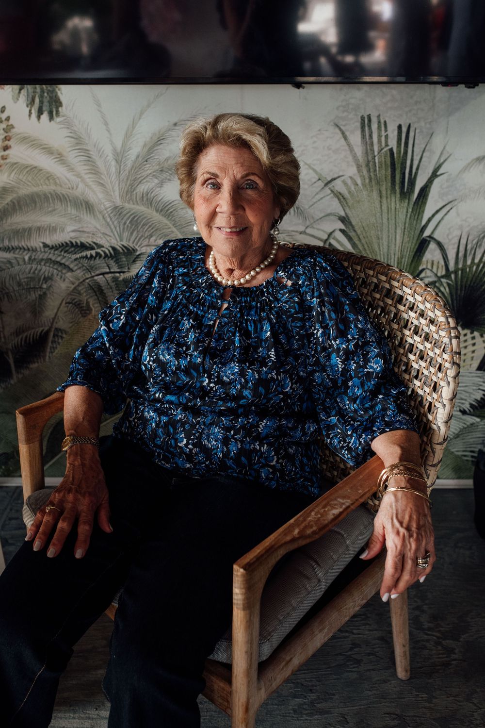 Elegant senior woman in blue floral blouse and pearl necklace seated in wicker chair at event.