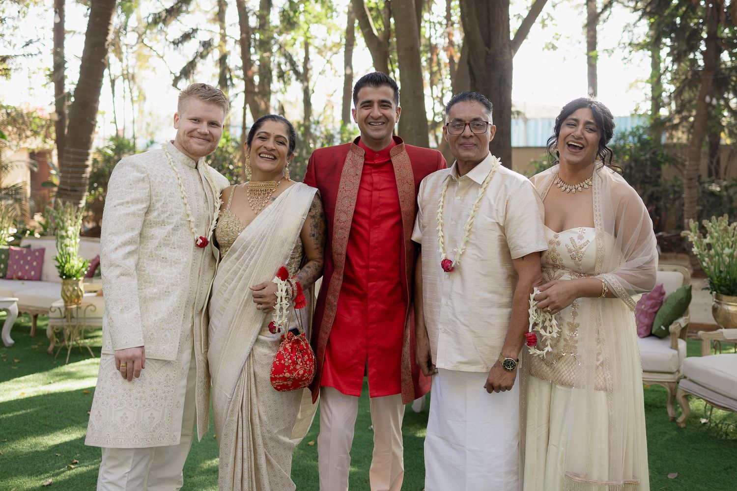 A group wearing traditional cream and red attire poses together in an outdoor garden setting.