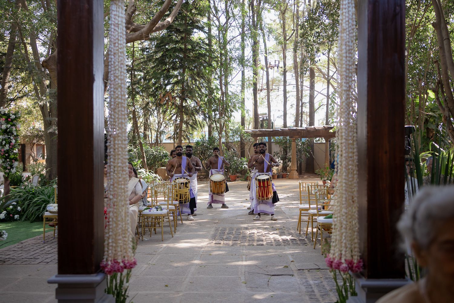 Outdoor restaurant seating area with wooden tables and chairs arranged under tall palm trees in a tropical setting.