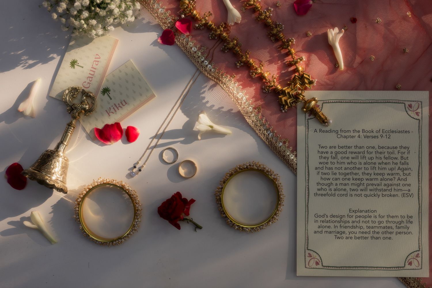 Wedding rings and red rose petals arranged on a white surface with scattered jewelry and decorative elements.