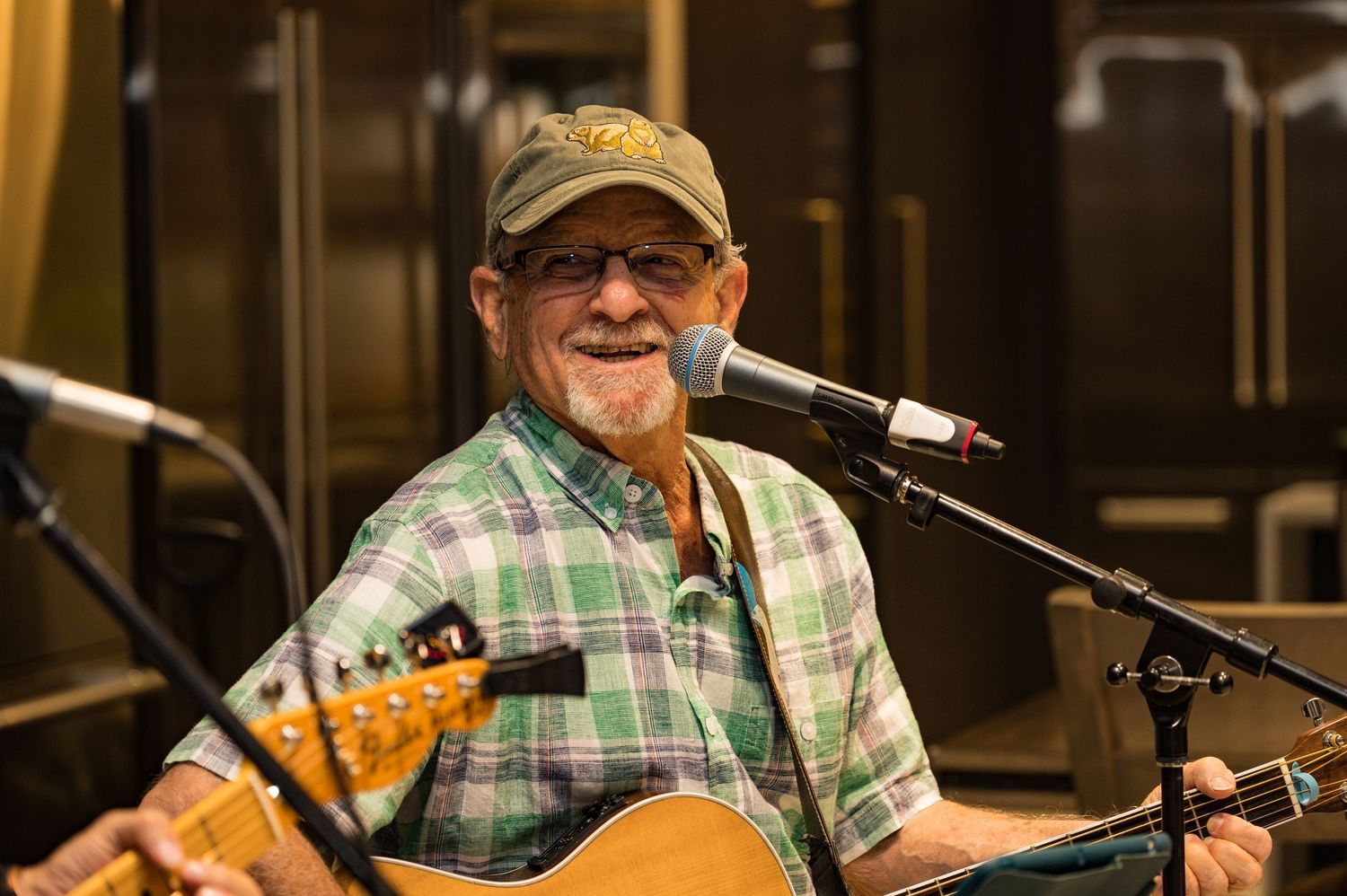 Smiling elderly man playing acoustic guitar and singing into a microphone at a live music performance.