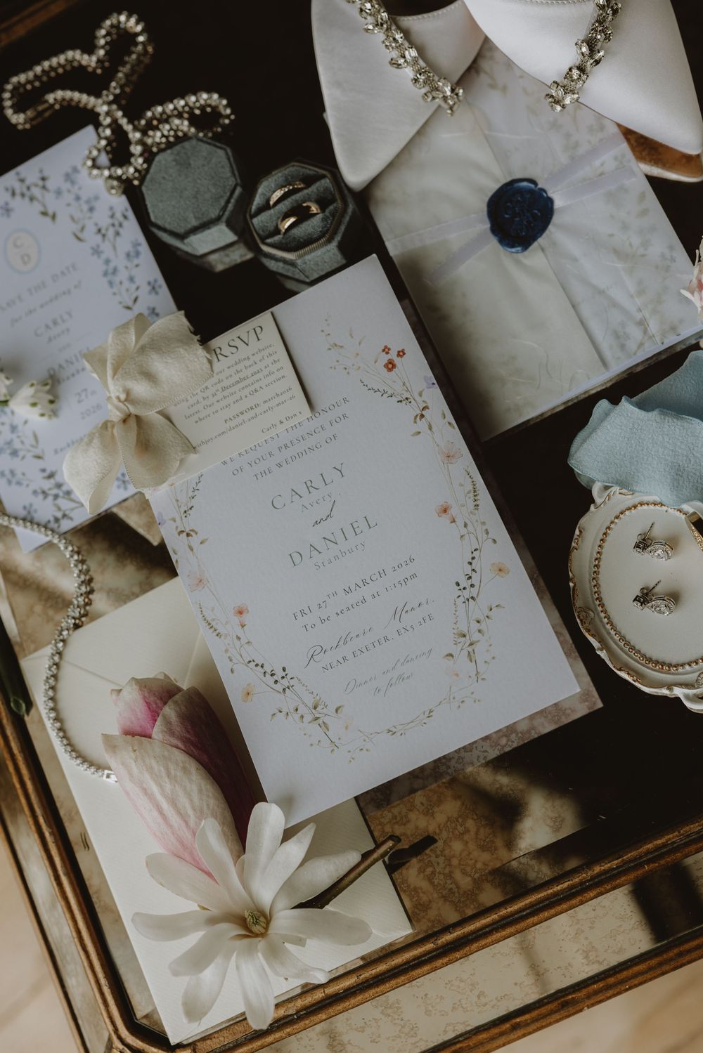 Elegant wedding invitation suite with floral details and accessories including pearls and white flowers on a dark surface.