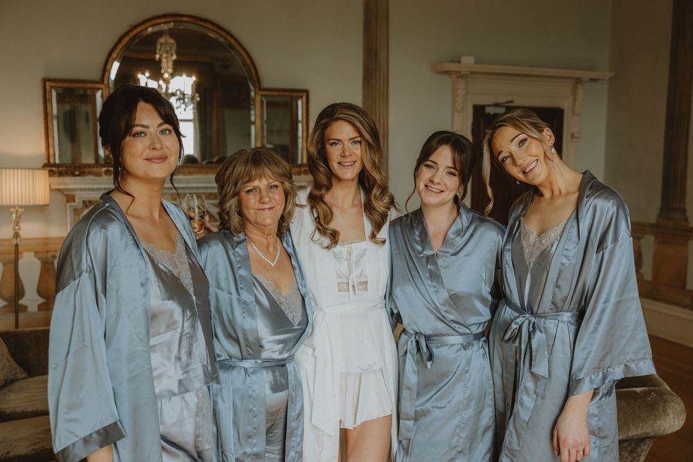 A bridal party poses together wearing matching blue silk robes while the bride wears white during wedding preparations.