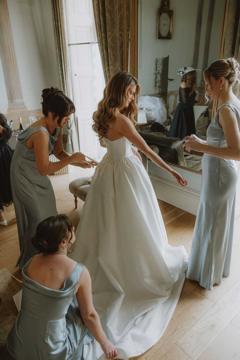 Bridesmaids in light blue dresses help adjust the bride's white wedding gown in an elegant room with natural light.
