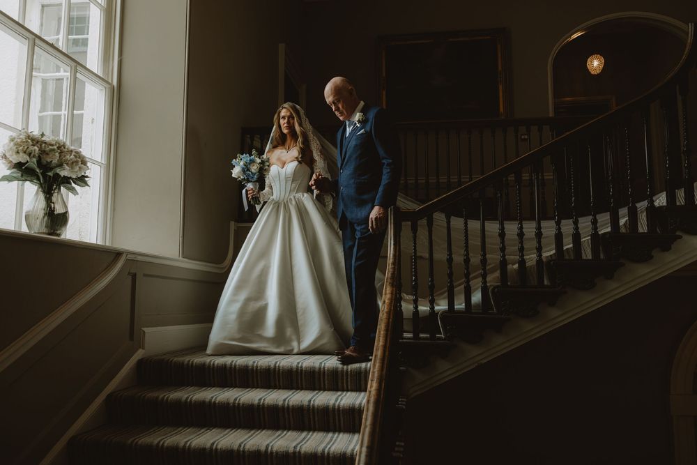 A couple stands on an elegant staircase with natural window light in a classic indoor venue setting.