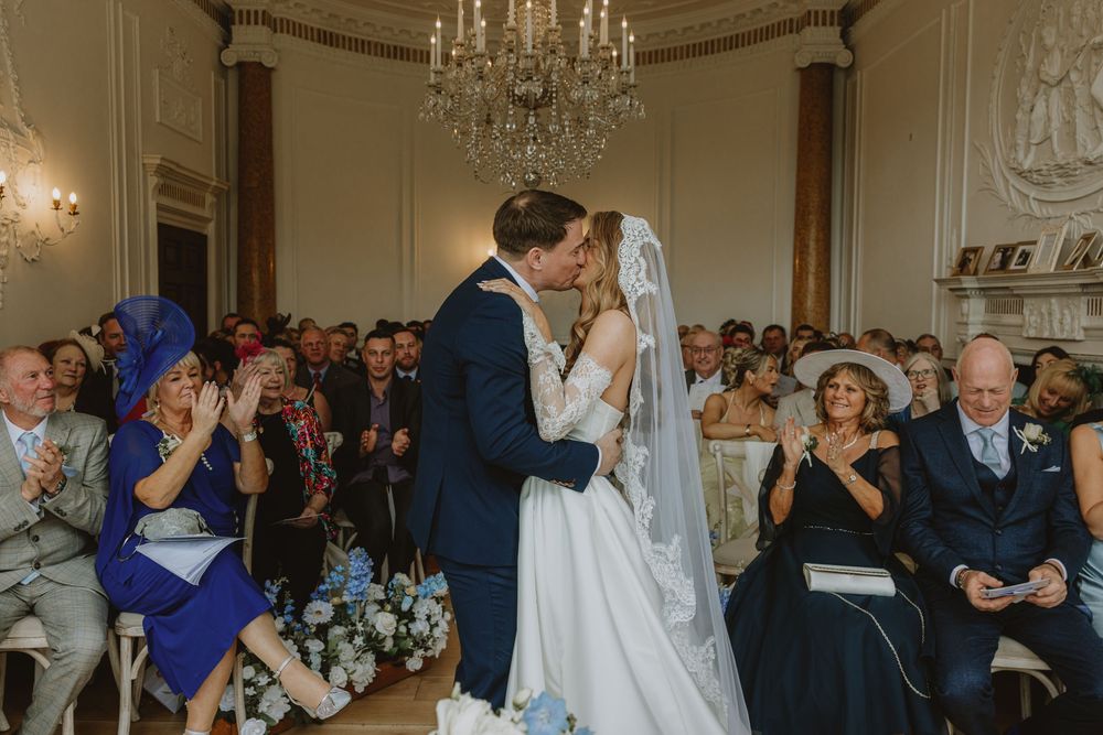 A couple shares their first kiss during a wedding ceremony under a crystal chandelier as guests look on.