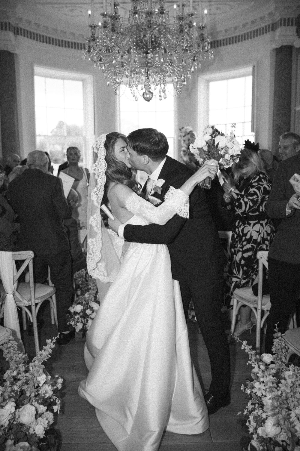 Wedding reception dance scene with guests celebrating under crystal chandeliers in an elegant white room.