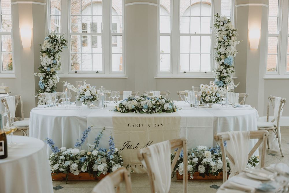 Elegant wedding reception table setup with white linens and floral arrangements in a bright room with tall windows.