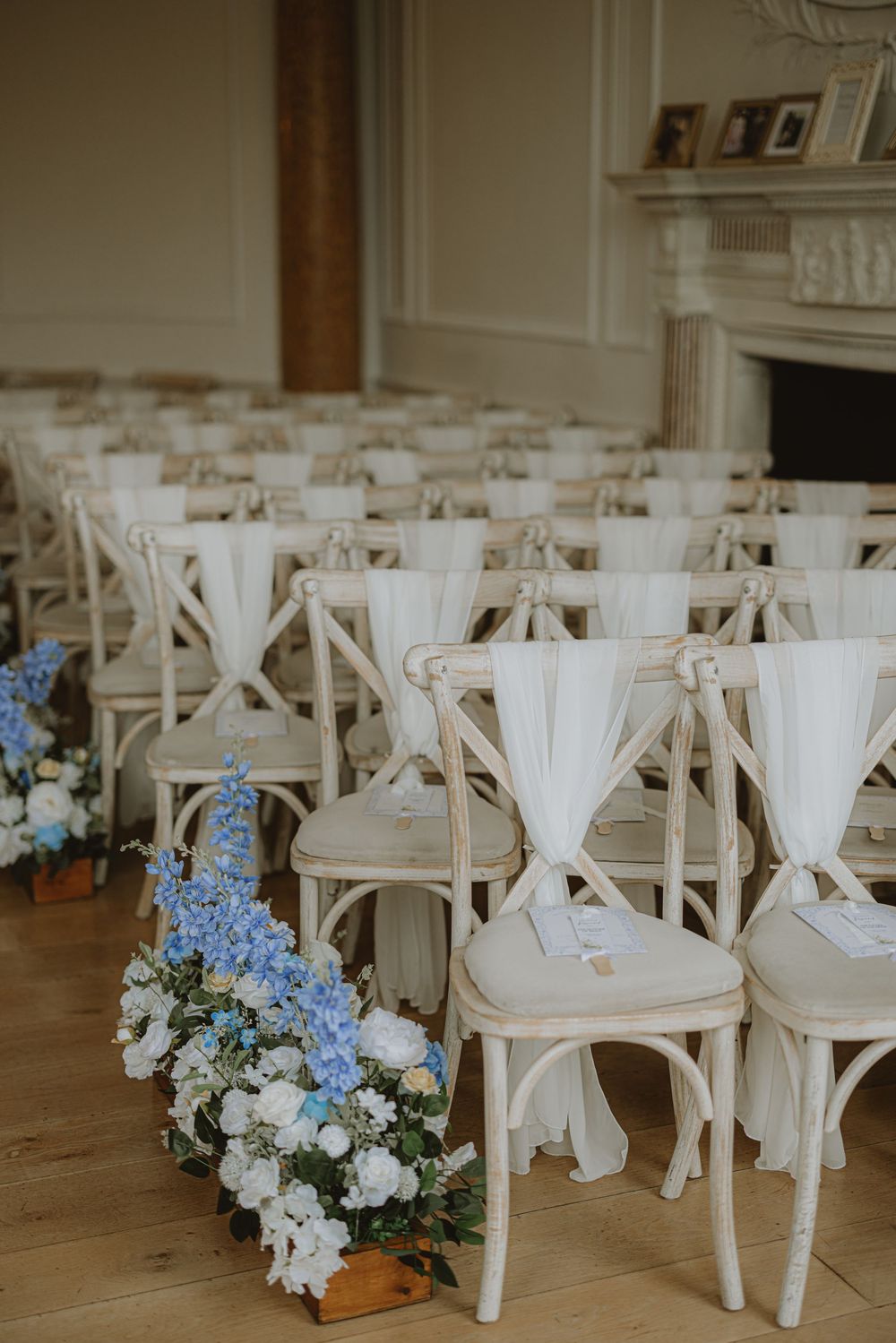 White cross-back chairs arranged for wedding ceremony with blue and white floral aisle decorations.