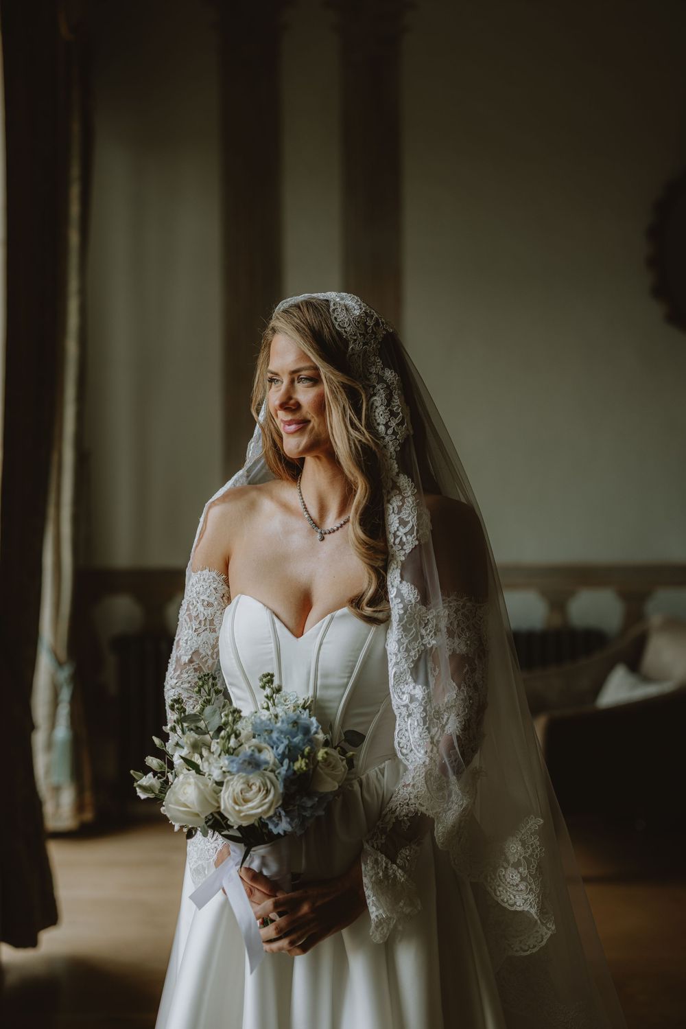 A bride in a strapless white wedding gown holds a blue and white bouquet while wearing an elegant lace veil.