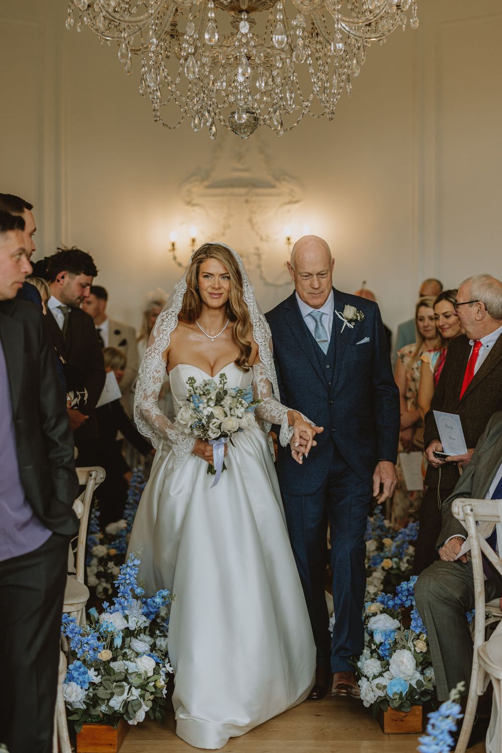 Wedding ceremony procession in an elegant room with crystal chandeliers and floral decorations.