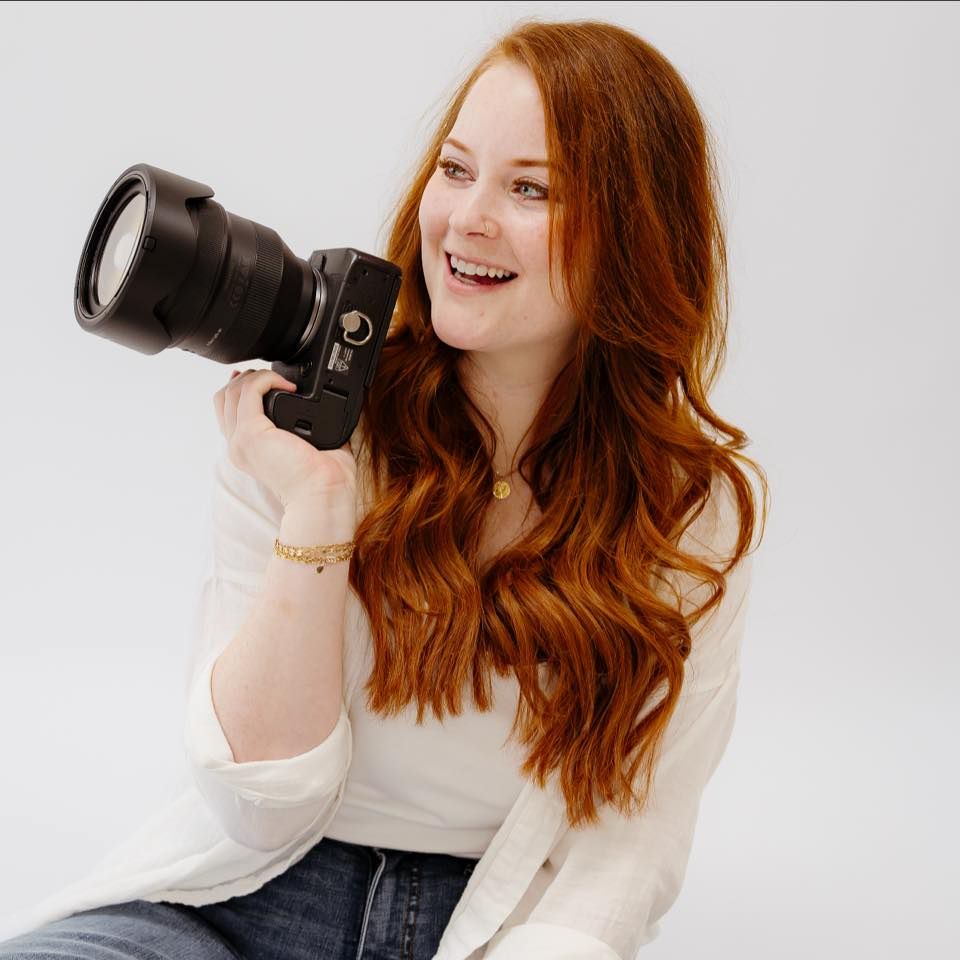 A smiling redhead woman holds a professional DSLR camera against a white background, wearing a white top and jeans.