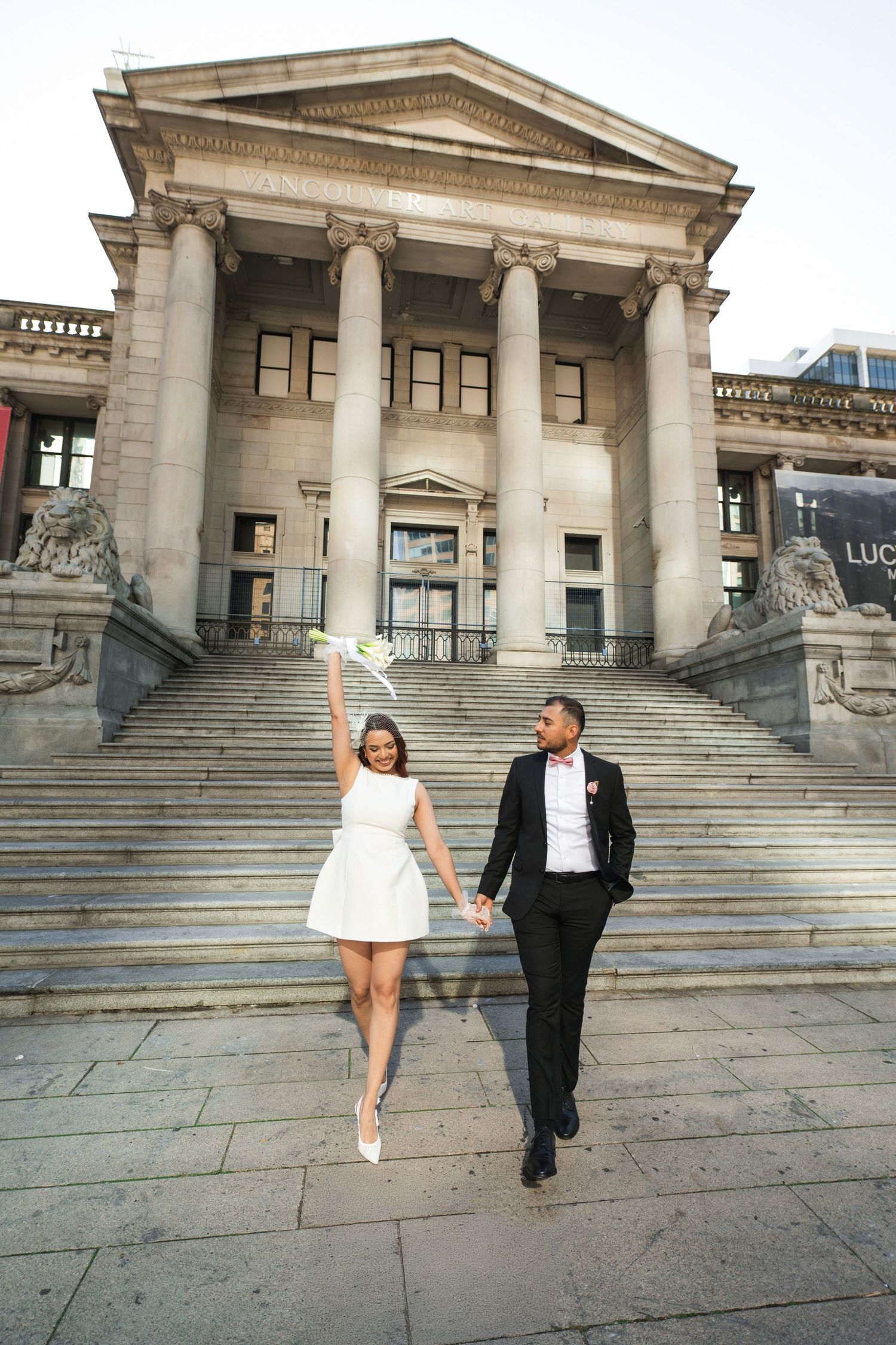 Bride and groom descend grand stone steps of classical building, bride raising bouquet in celebration.