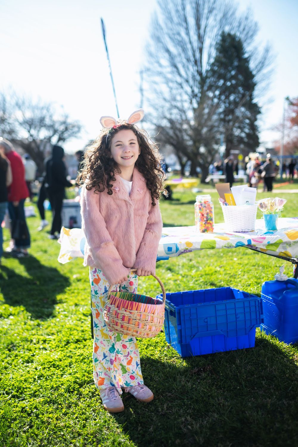 Young girl in pink fur coat and bunny ears holds Easter basket at outdoor egg hunt event.