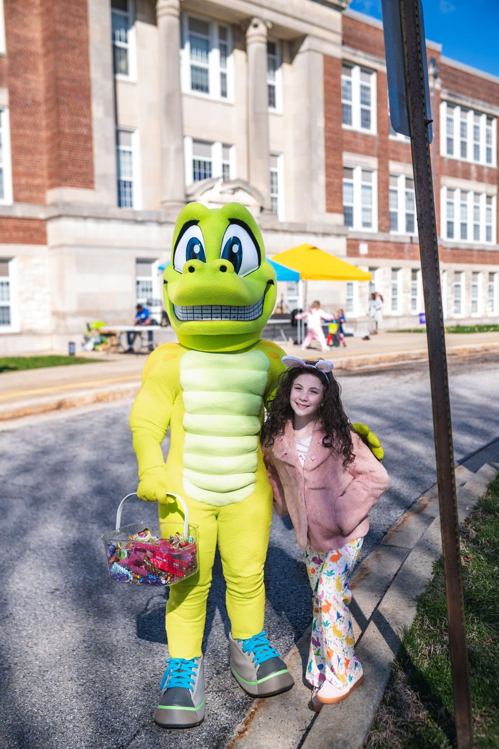 A young girl poses with a yellow dinosaur mascot holding flowers outside a brick school building.