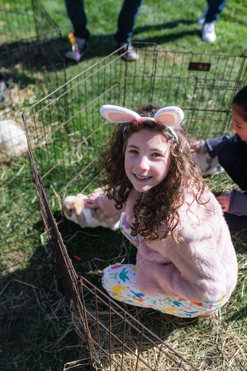 A smiling girl wearing bunny ears crouches by a wire pen petting a small animal outdoors at an Easter event.