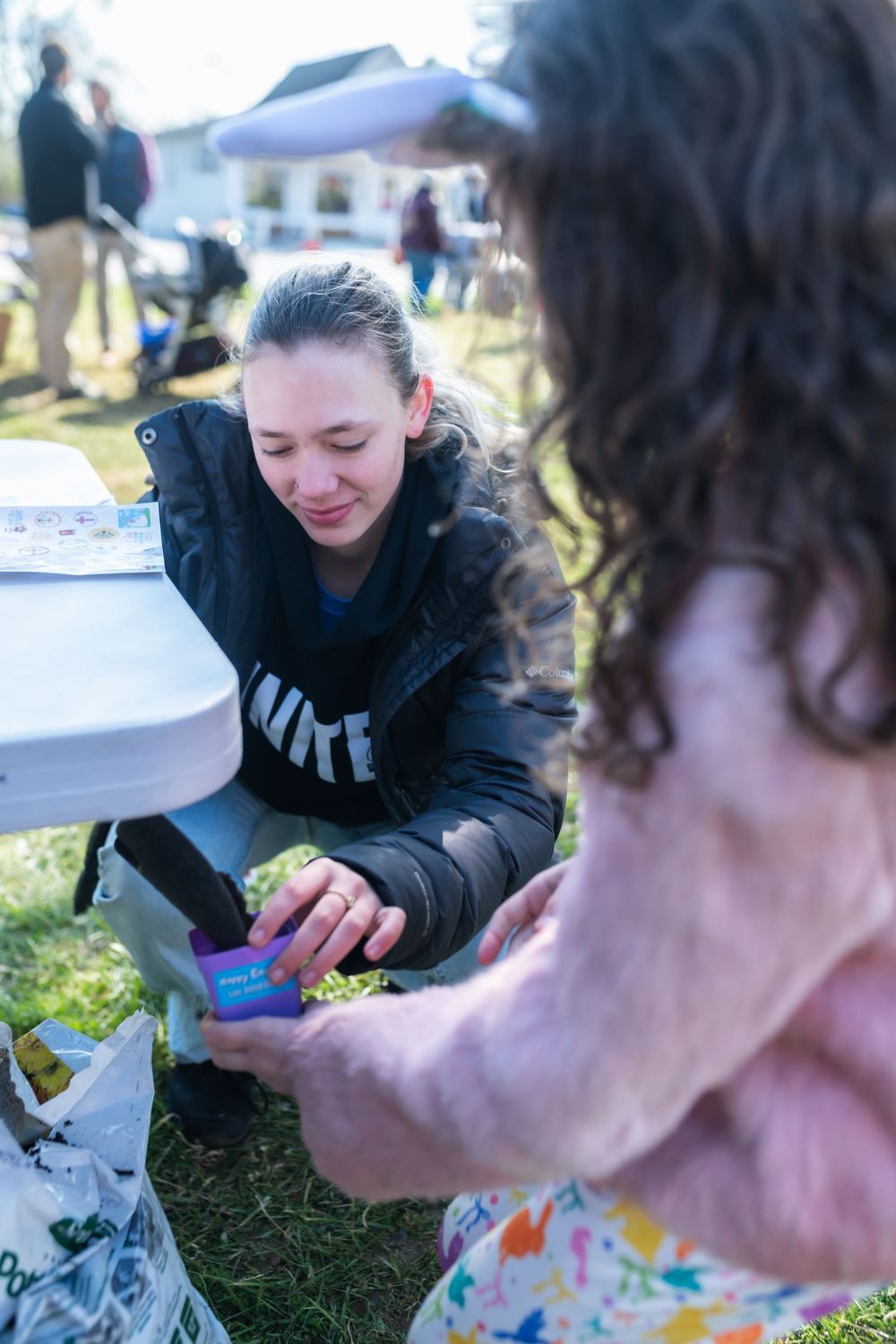 A woman in a Nike jacket hands a purple gift to a curly-haired girl at an outdoor community event.