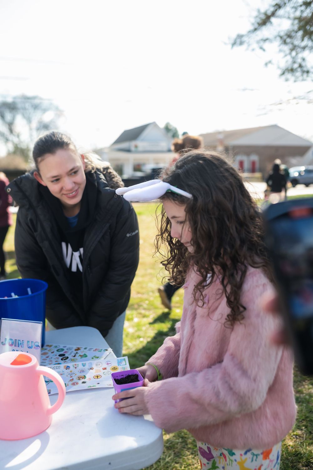 A young girl in a pink coat and bunny ears examines a purple toy at an outdoor spring event table.