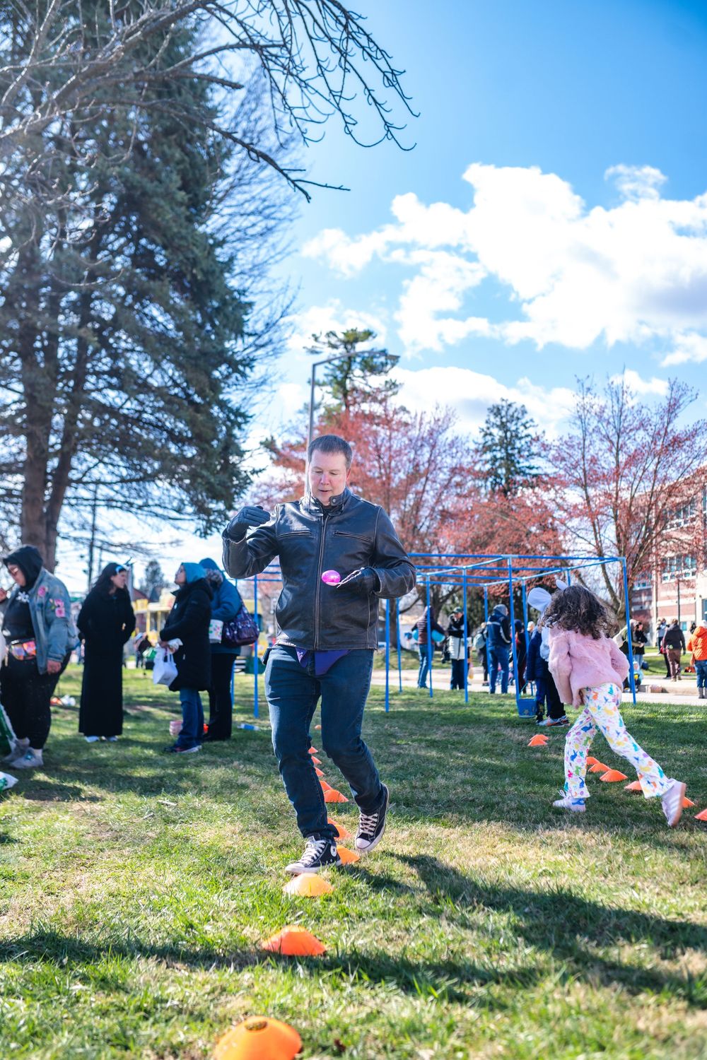 Children and adults enjoy an outdoor Easter egg hunt on a sunny day in a grassy park with trees and playground equipment.
