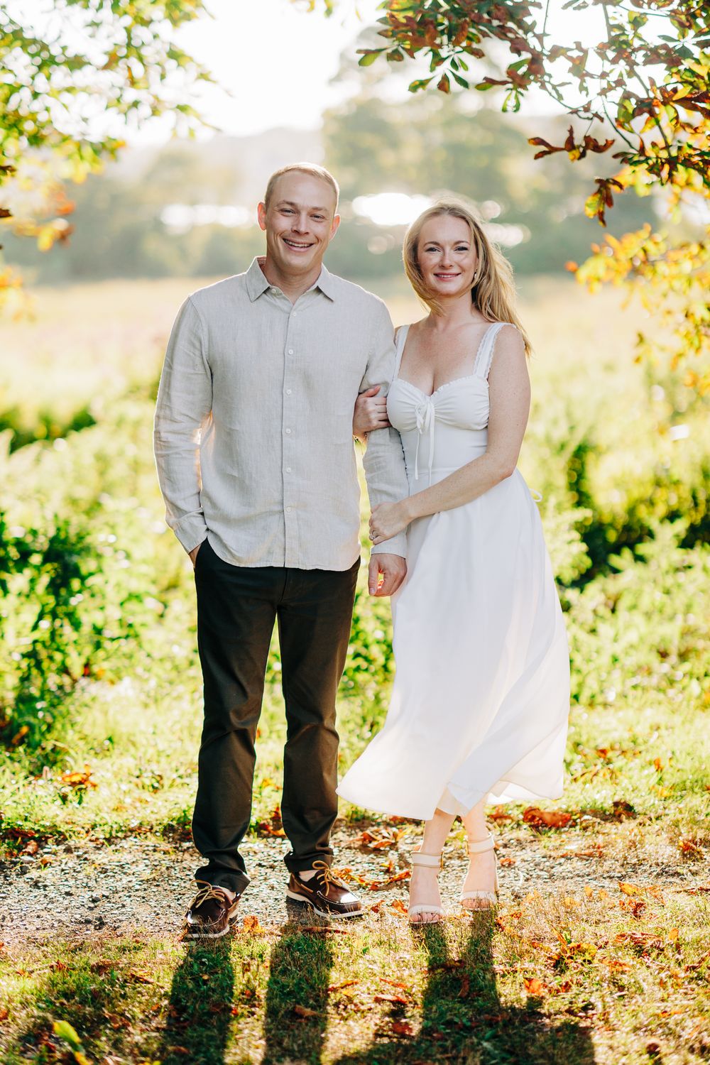 A couple in white and grey attire poses playfully in a sunlit garden setting during an autumn photo session.