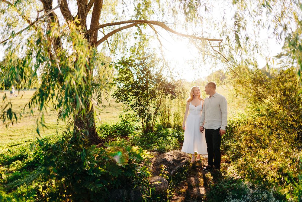A couple walking through a sunlit garden path with overhanging trees creating a romantic natural archway.