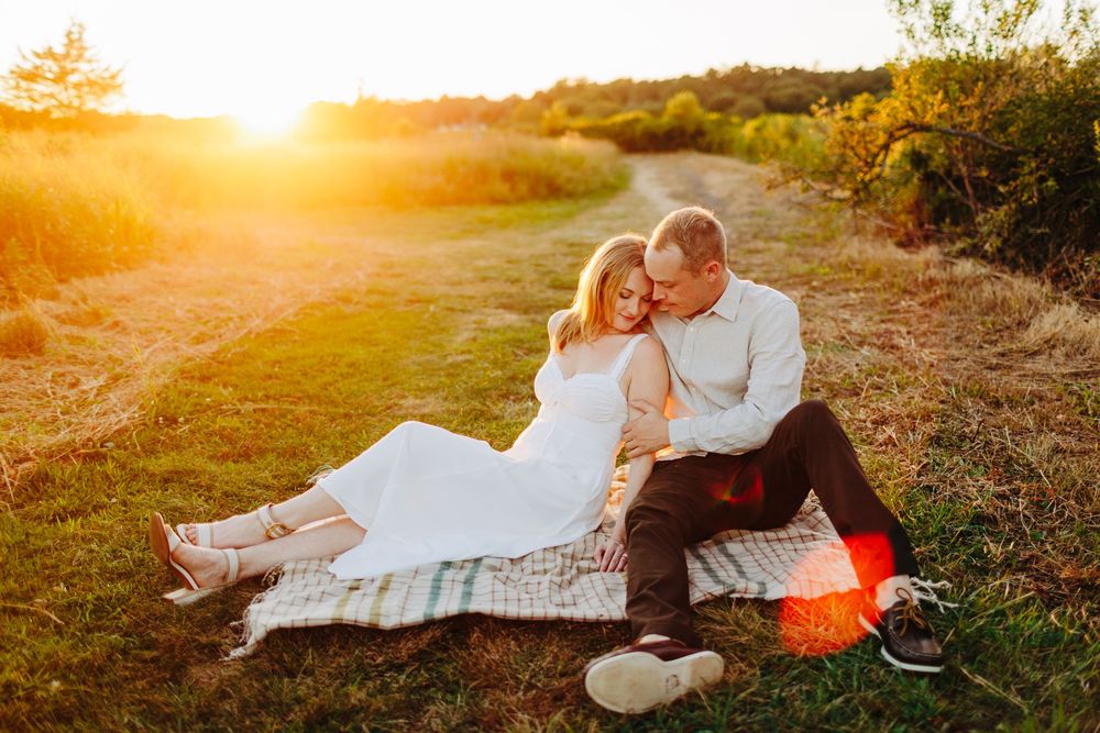 A romantic couple sits on a blanket in a grassy field at golden hour as the sun sets behind them.