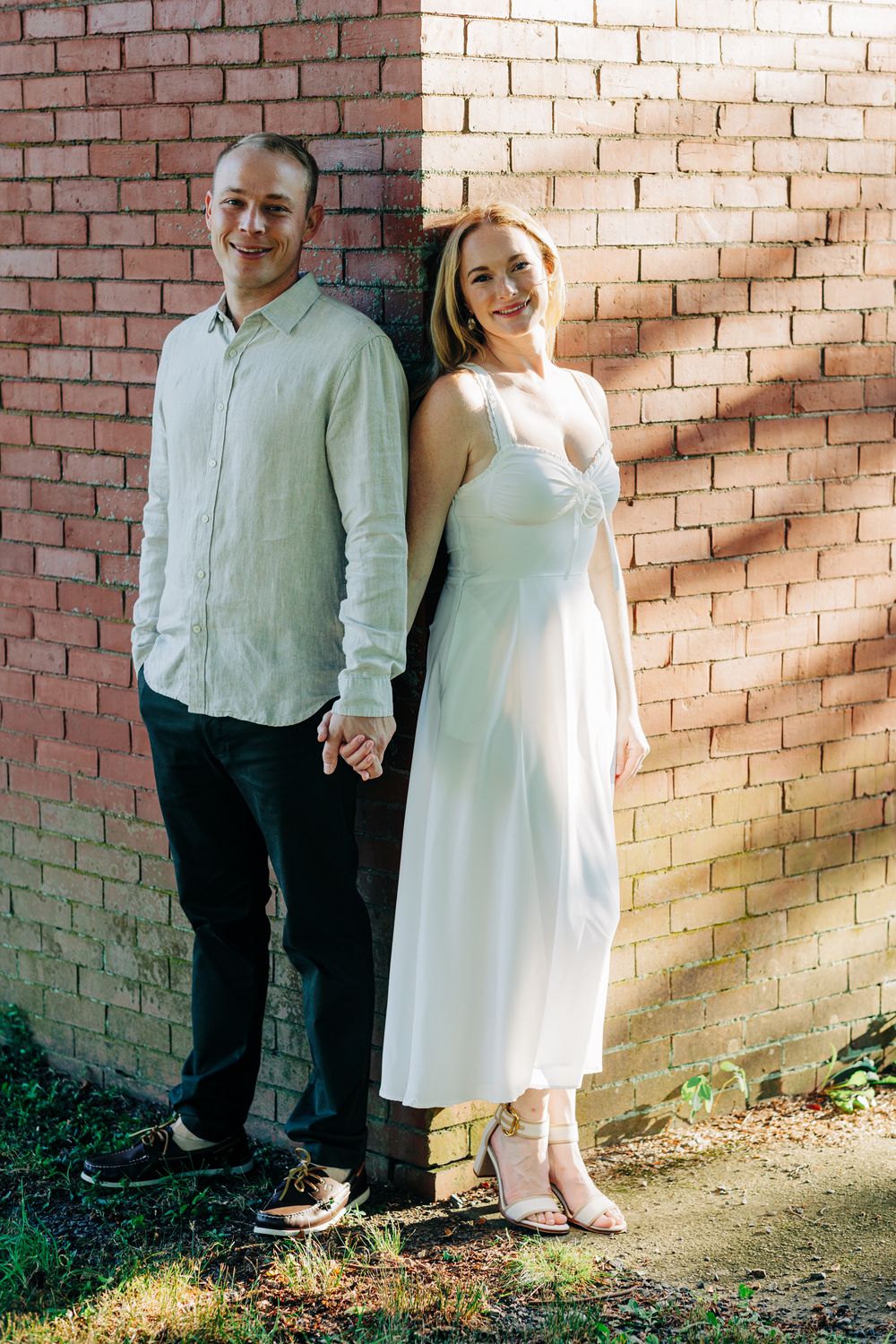 A couple in coordinating white outfits poses against a brick wall in warm sunlight for romantic photos.