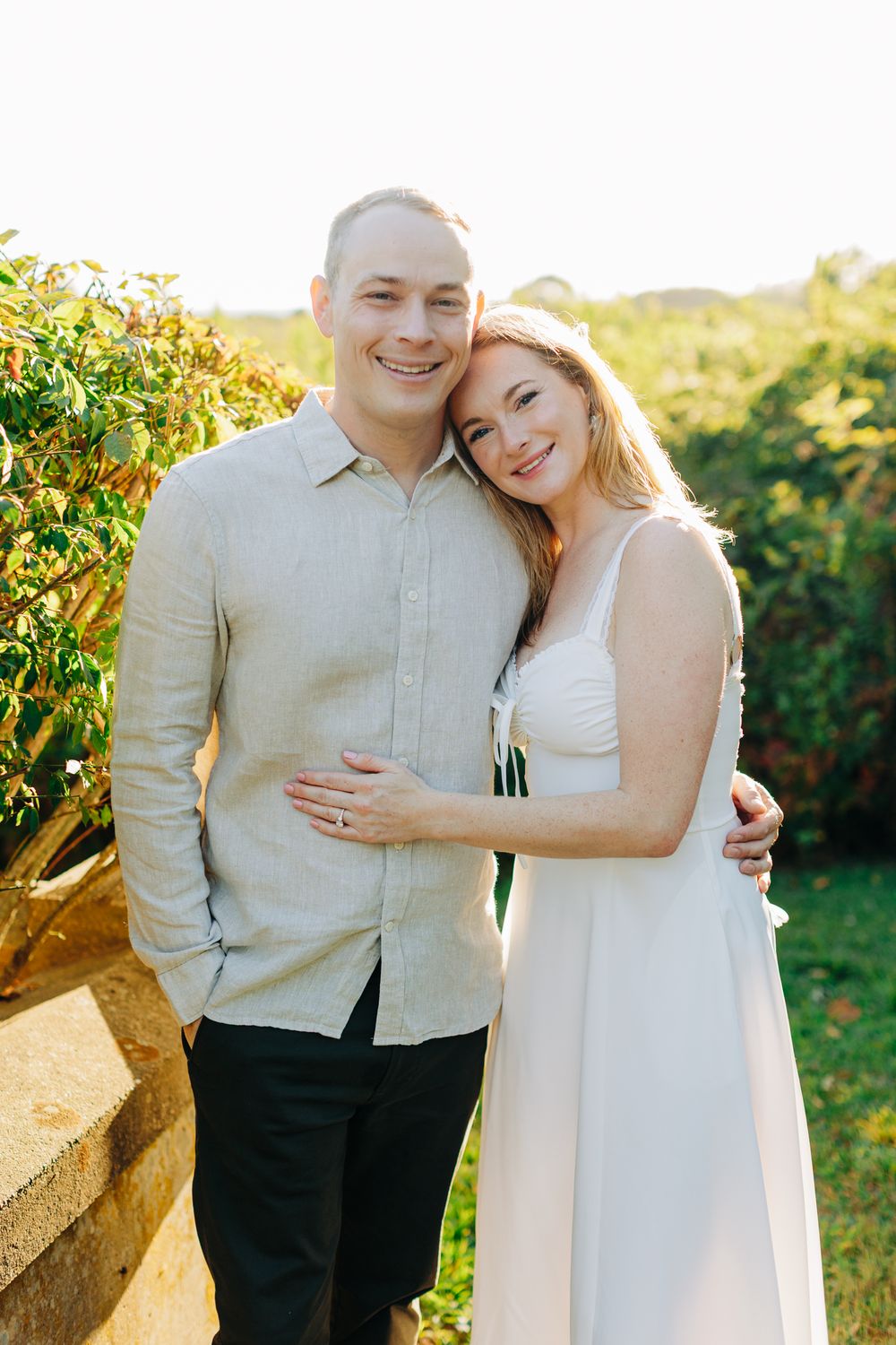A couple embraces outdoors during a sunset photoshoot wearing light colored clothing against a natural backdrop.