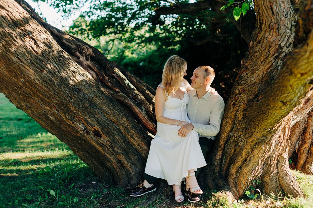 A couple sits together on a large tree trunk in a sunny garden setting wearing light colored casual clothing.