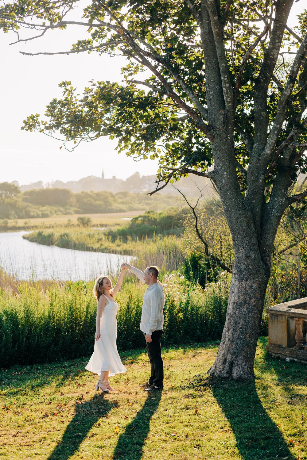 Two figures stand beneath a large tree by a serene lake at sunset, casting long shadows on the grassy shore.