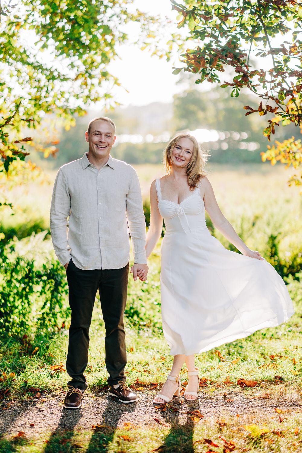 A couple poses together in a sun-drenched garden setting, with the woman wearing a flowing white dress and holding its hem.