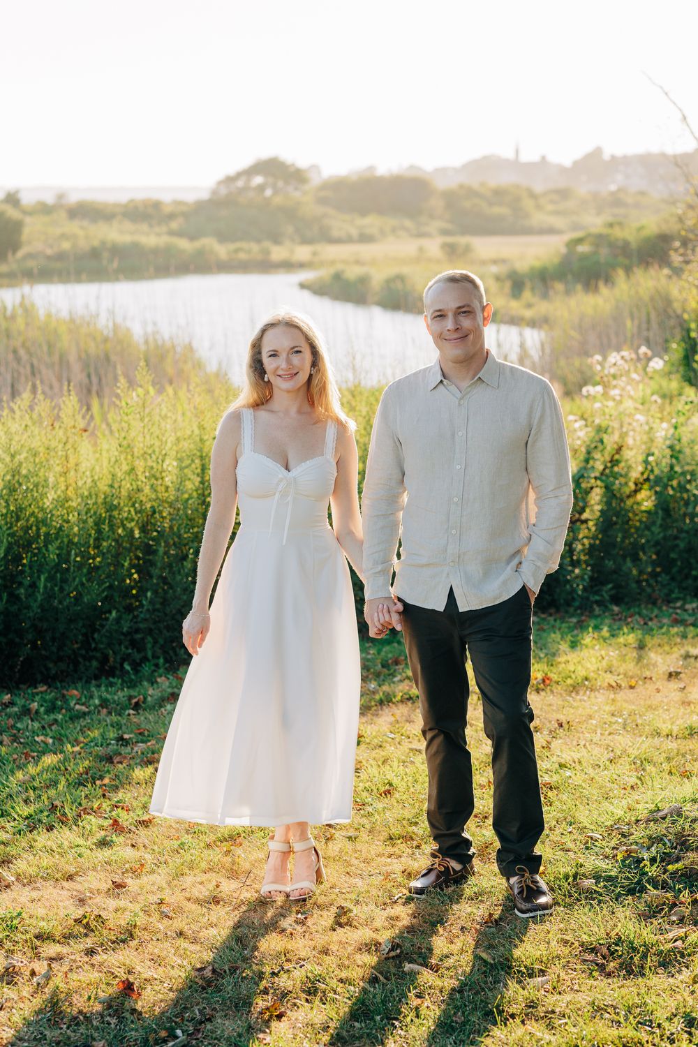 A couple stands together in a sunlit meadow casting long shadows during golden hour.