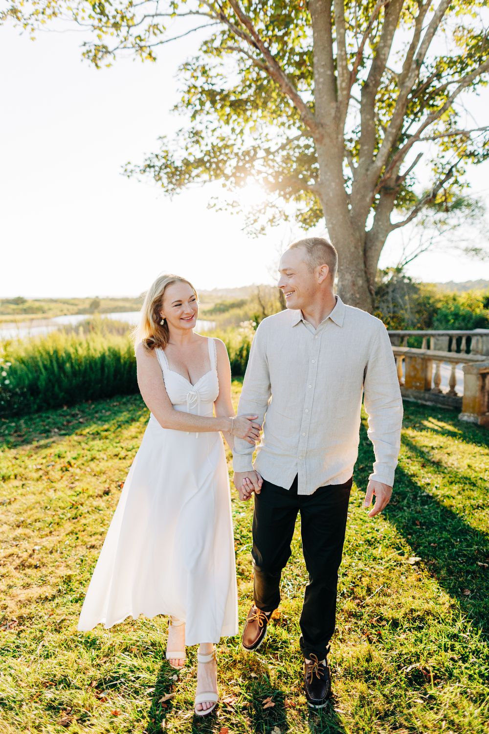 A couple in white and black attire walks hand in hand through a sunlit grassy field during golden hour.