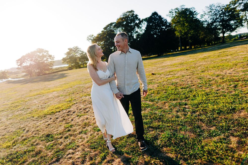 A couple walks hand in hand through a sunlit meadow during golden hour at sunset.