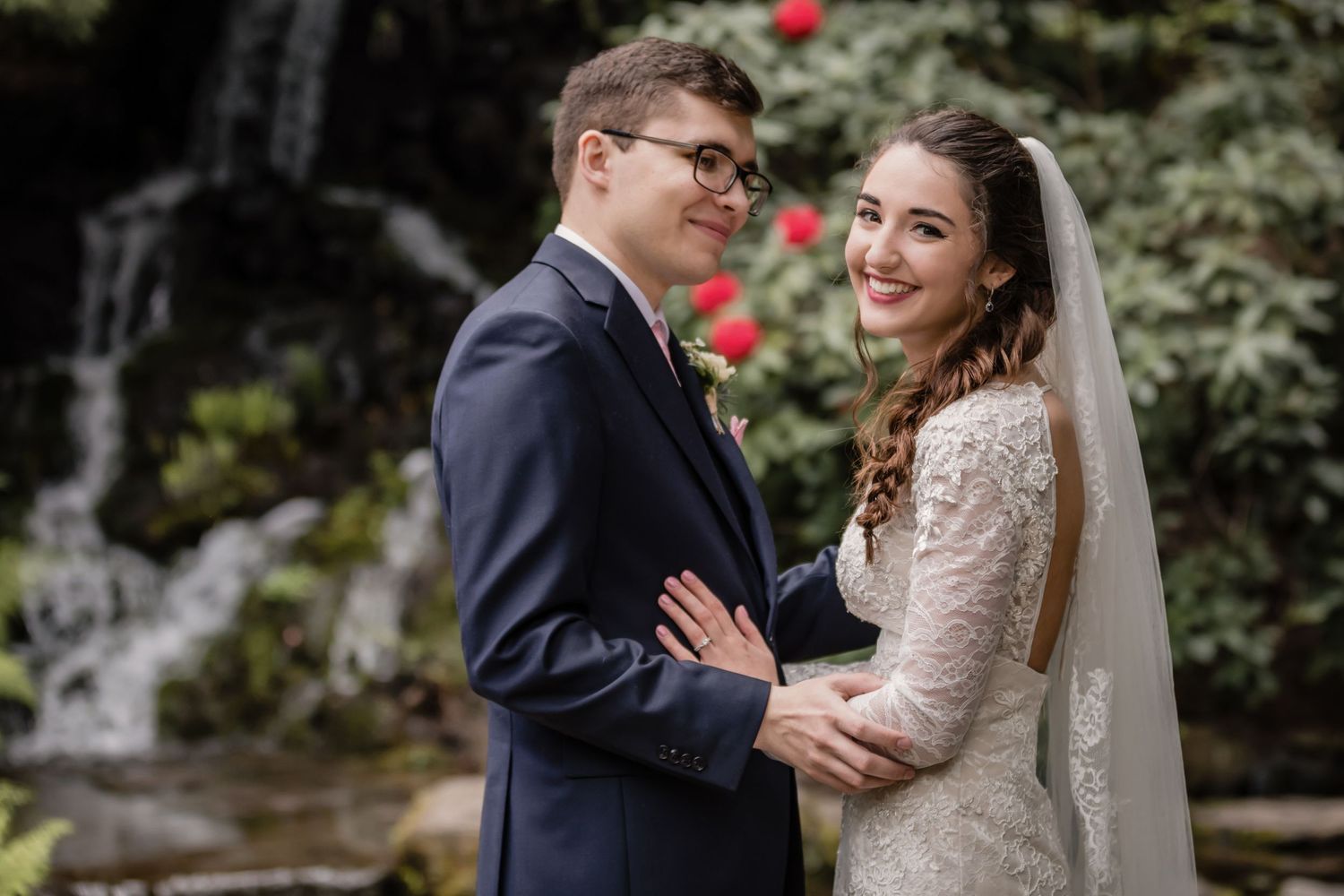 A romantic wedding couple shares intimate moments in a lush garden setting with red flowers in the background.