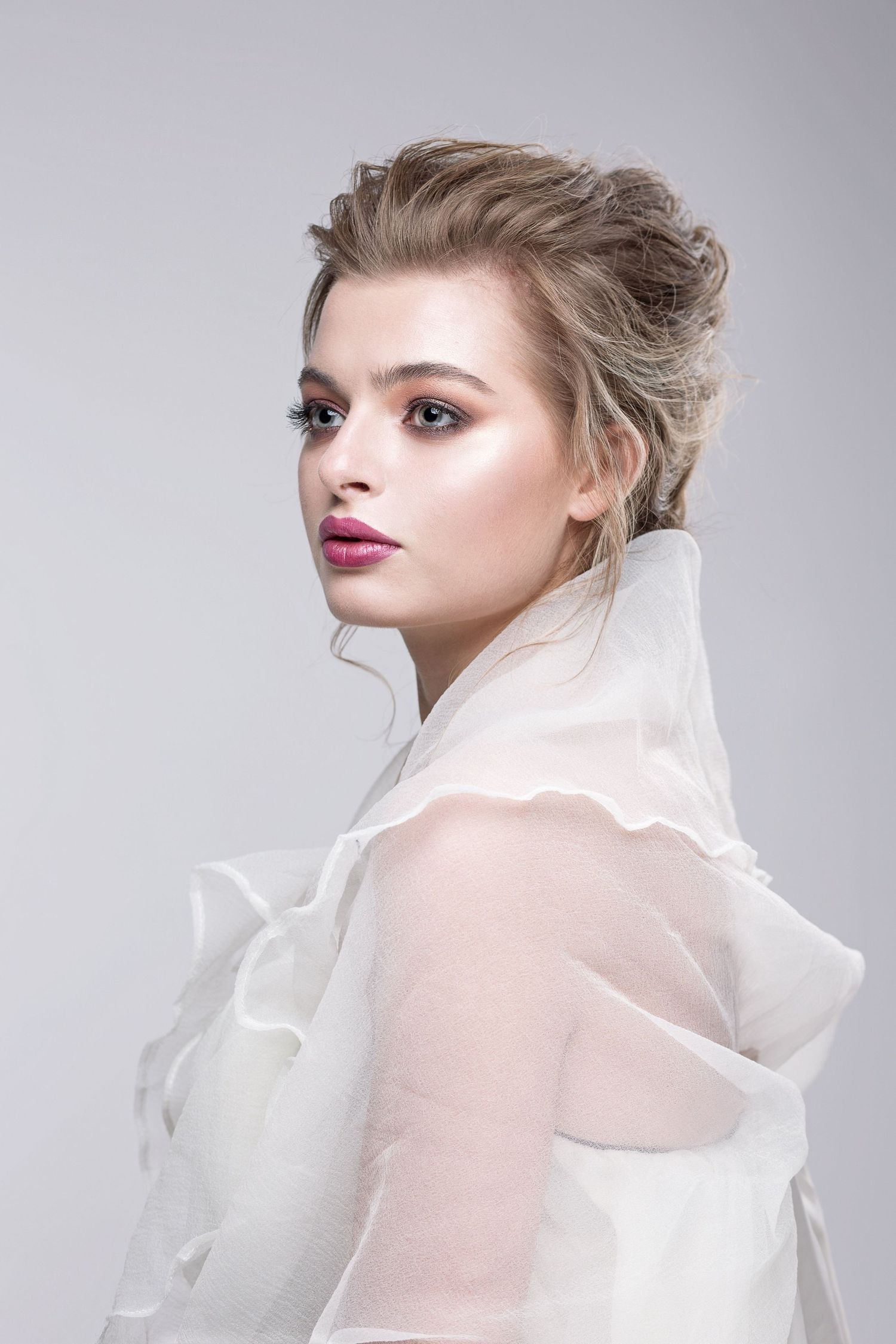 Fashion portrait featuring ethereal sheer white fabric and dramatic makeup styling against a light gray backdrop.