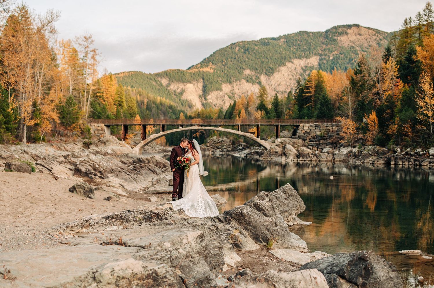 Bride and groom embrace on rocky riverbank with autumn trees and stone arch bridge in mountain landscape.