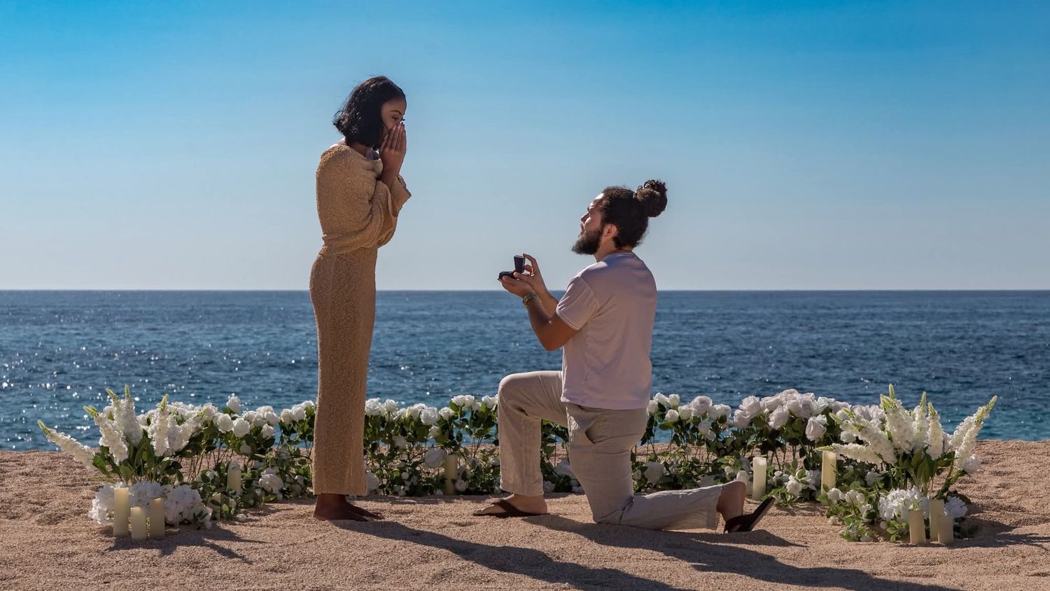 Man proposes to woman on one knee by the ocean, surrounded by white floral arrangements on a sunny beach.